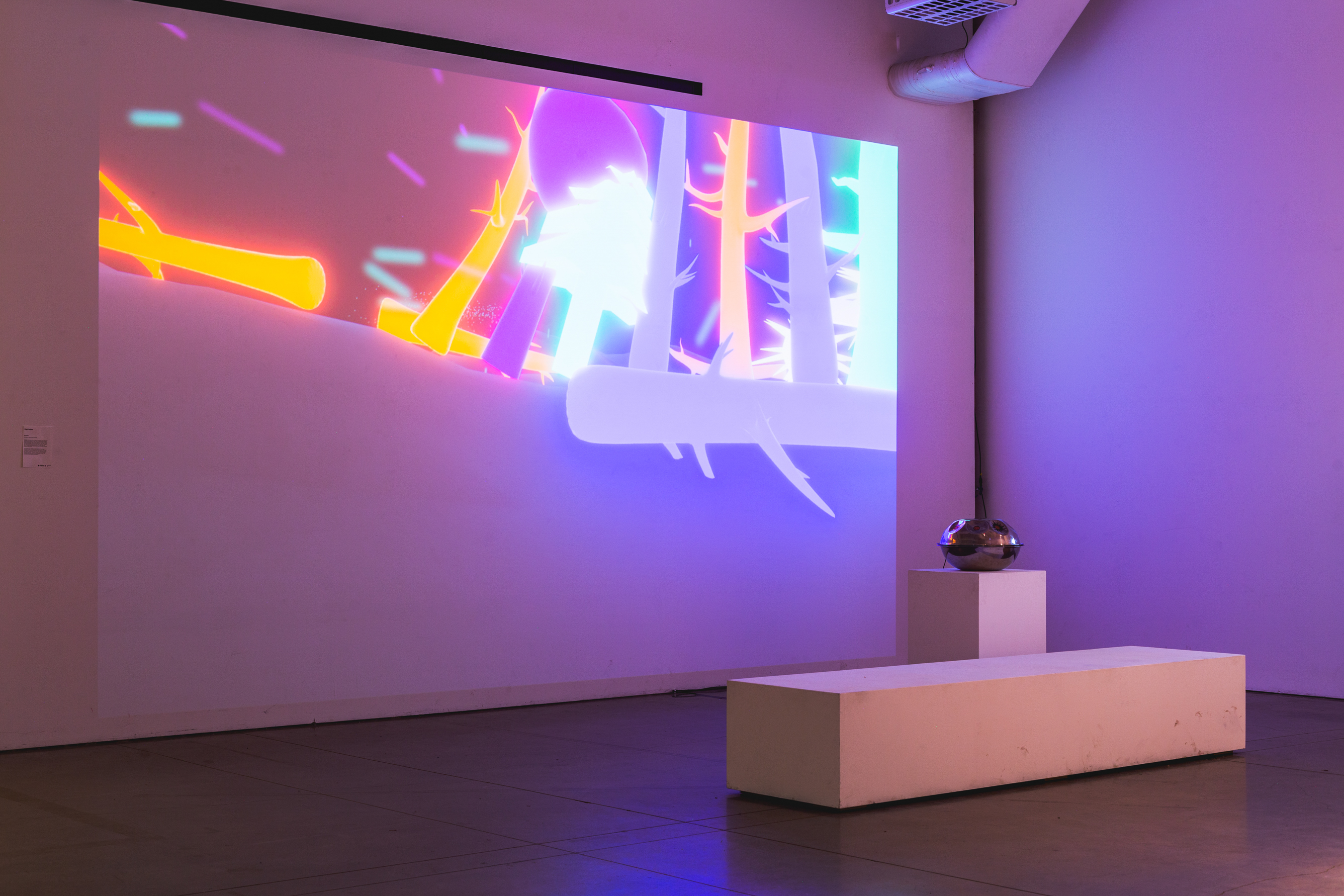 A projection onto a large white wall, the projection is an interactive 3D game. In front of the projection is a wide white bench and a musical instrument on a white podium.